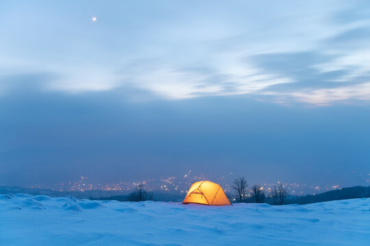 Yellow Tent Lighted From The Inside Against The Backdrop Of Glowing City Lights In Fog. Amazing Snowy Landscape. Tourists Camp In Winter Mountains. Travel Concept