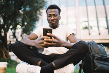 Pensive dark skinned male student in eyewear for vision correction sitting outdoors on street with backpack and mobile phone, serious african american hipster guy blogger checking mails on smartphone