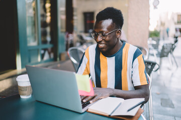 Obraz premium Dark skinned male user in spectacles installing application for creating blog publication via laptop technology, cheerful hipster guy searching content information while browsing wireless on netbook