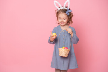 Funny child girl in Easter bunny ears holding basket with colorful eggs on pink background