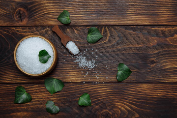 White sea salt in a bowl with a spoon and green ivy leaves, top view flat composition with empty space for text on brown wooden background, spa treatment concept, bath and sauna, natural scrub