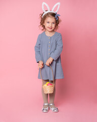 Funny child girl in Easter bunny ears holding basket with colorful eggs on pink background