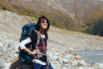 Young woman with a backpack in the mountains sits on a stone, resting and looking at the mountain view