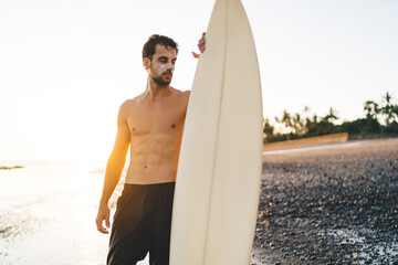 Fit surfer standing on pebble beach with surfboard in sunlight