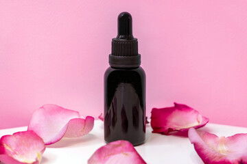 Black bottle of essential oil with rose petals around it on pink background