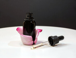 Opened black bottle of essential oil with dropper full of oil. Rose petals around flacon. Black and white background