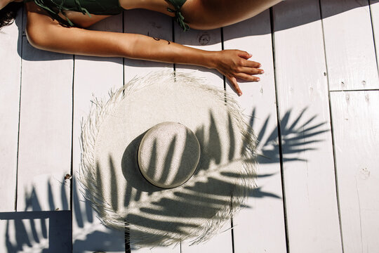 White hat on white wooden planks by the pool under the shade of a palm branch. A woman's body in a swimsuit is slightly visible