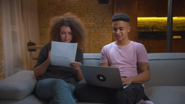 Young mixed race couple cheating while online interview for new job sitting on sofa. African American man giving high five to girlfriend for exam passing help.