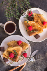 thin homemade pancakes with raspberries and chocolate on a dark background on a light plate, with herbs and tea, close-up, vertical arrangement, top view