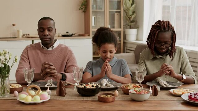 Medium close-up of happy Black family sitting at table with eyes closed, praying before Easter dinner, then father, daughter and mother taking plates with food