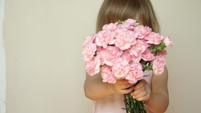 Portrait of little child girl holds bouquet of flowers, pink carnations, looks at camera, laughs, smiles and smells flowers. Spring, summer concept
