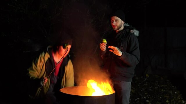 Two homeless young men are frying apples on a fire. Men stand at night near a barrel of fire and bask.