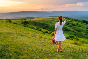 Holiday in the country with a Caucasian woman dressed in a white dress sitting barefoot in the grass and enjoying the beautiful landscape of nature with green hills and dense forest at sunset.