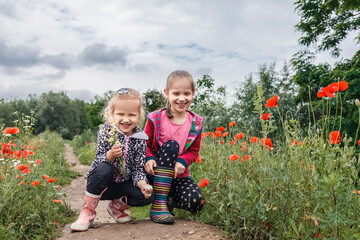 Two little girls on a background of a field of poppies and trees. Two children in rubber boots with flowers in their hands