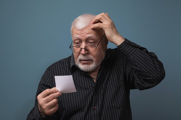 an elderly man sits in glasses, choking his head in surprise, looks at a sheet of paper he holds in...