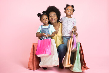 Portrait of a young attractive African American mother and her two cute little daughters, posing to camera over isolated pink background with a lot of colorful shopping bags