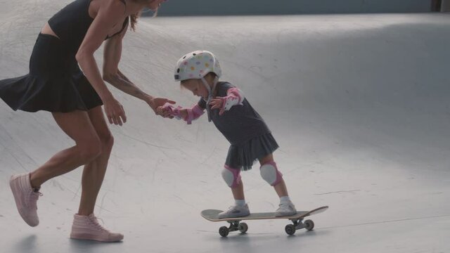 Real time of young Caucasian woman wearing streetwear holding hand of her little daughter wearing safety helmet, knee and elbow pads and helping her skateboarding in skate park