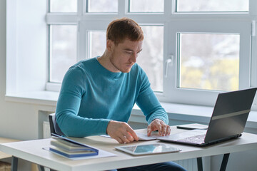 Smiling handsome young guy sitting at table, looking at computer screen. Focused millennial man working in modern office or studying online at home.