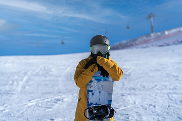 Fototapeta na wymiar A snowboarder in a yellow jacket poses on a mountain with his arms crossed over a snowboard.The cable car can be seen in the blurred background