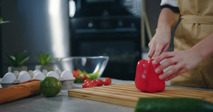 Healthy nutrition concept where unknown woman cutting on half bulgarian pepper with knife on cutting board when preparing vegetable salad in kitchen