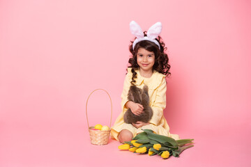 child girl in bunny ears holding rabbit, tulips, basket with colorful eggs on pink background.