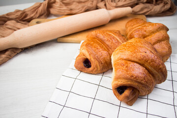 Food photo. Three tasty croissants lay on linen fabric near wooden rolling pin on white table. Rustic style