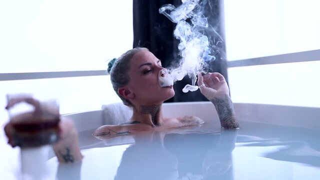 A young sexy tattooed girl enjoying a cigar and a drink while having a bath in a relaxed atmosphere in the bathroom with a bunch of money in the water. Relaxation, luxury, bathroom, home