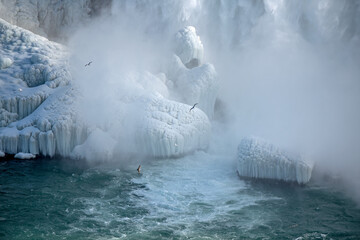 Freezing mist creating ice formations at the bottom of Niagara Falls, Ontario, Canada. Seagulls fly through the mist above the river.