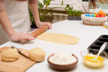the process of rolling out the dough at home on the table is the hands of a woman for making cruffins festive pastries for Easter . side view of a bright kitchen , with painted eggs