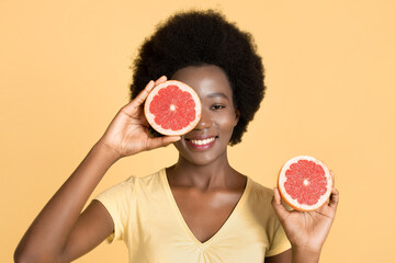 Close-up portrait of charming african girl holding halfs of grapefruit, hiding her eye. Amazing smiling dark skinned woman with grapefruit slices looking at camera on yellow background