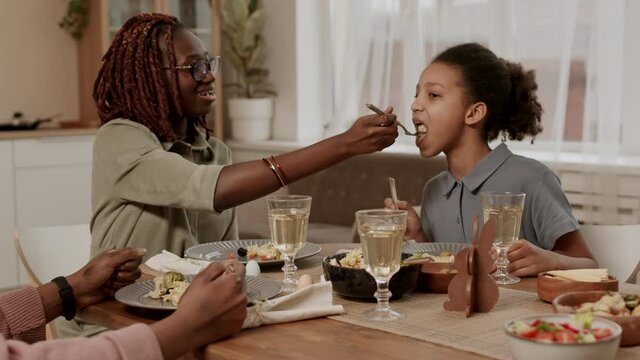 Chest-up of young cheerful African woman sitting at table, having family festive dinner, sharing some of her food with daughter feeding her off fork, they smiling