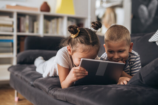 children are playing together at home. They are using digital tablet while laying on a sofa.