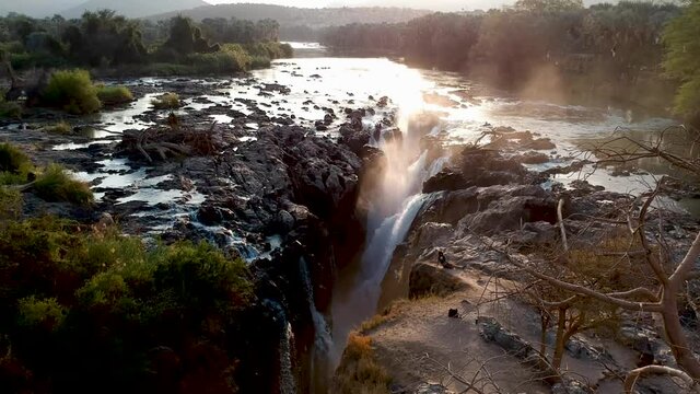 Panning down into Epupa falls with big Baobab and the mst from the waterfall with sun setting on the water of the Kunene river Aerial