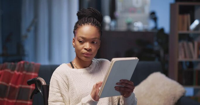 Young beautiful african business woman in wheelchair using mobile apps on tablet computer screen freelancing from home. Apartment. Girl with disabilities.