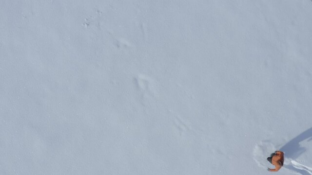 Top-down aerial view of a middle aged man wearing an orange winter jacket making a snow angel