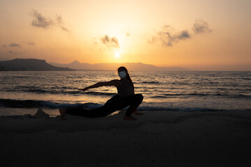 Fototapeta premium Silhouette of unrecognized young woman with protective surgical face mask performs yoga stretching exercises at the beach at dusk during coronavirus pandemic.