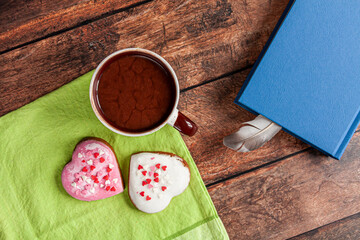 a cup of hot chocolate and two heart-shaped gingerbreads on a green towel ans a thick old-fashioned brown book with a feather on brown wooden background