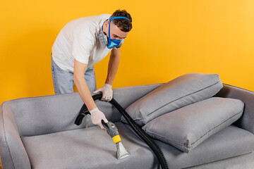 Young male worker cleaning sofa with vacuum cleaner.