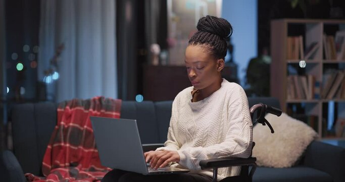 Indoor portrait of young beautiful african woman holding laptop sitting in wheelchair smiling to camera. Apartment interior. Ethnicity. Disabled person.