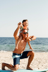 papà con occhiali da sole in costume da bagno insegna le basi del surf alla figlia in una spiaggia...