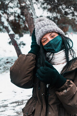Woman portrait, she is happy in the snow winter forest, wears a mask for covid protection beanie in the head gloves  to the hands and scarf in the neck