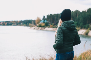 A young blonde woman in a dark green jacket stands on the edge of a cliff and looks into the distance against the background of a forest landscape. Absence, loneliness, depression, thoughts about life