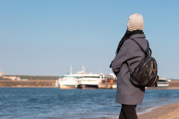 A young brunette woman in a gray coat stands on the beach and looks out at the ships in the...