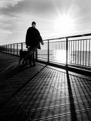 Czarno-białe pionowe ujęcie mężczyzny w maseczce spacerującego z psem po kładce dla pieszych w Kołobrzegu