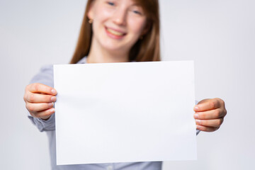 Fototapeta premium Woman with a piece of blank, empty space, paper sheet in her hand for your ad. Surprised woman shows a poster, canvas, white sheet of paper isolated on gray background