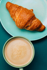 A teal cup with coffee on a blue background next to a croissant. Everything have the aquamarine color. Spring breakfast 