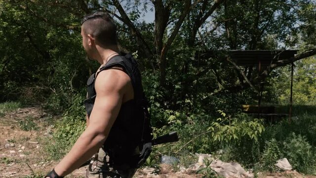 A muscular rebel in military pants and an unloading vest walks through an abandoned area with a machine gun in his hands