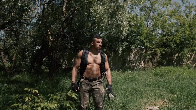 Unflappable gunman in a protective vest, handcuffs hanging on it and rifle in hands walks through a deserted garbage-strewn territory