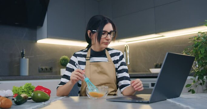 Pleasant concentrated 30-aged bruneete in glasses sitting in front of laptop and searching recipe while mixing eggs in transparent bowl with whisk