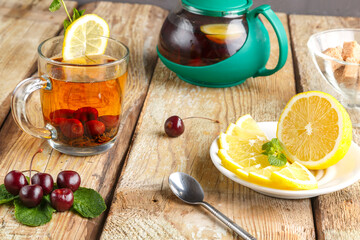 black tea in a glass cup with mint cherries and lemon on a wooden table next to fresh cherries and a teapot and lemon in a plate and mint leaves.